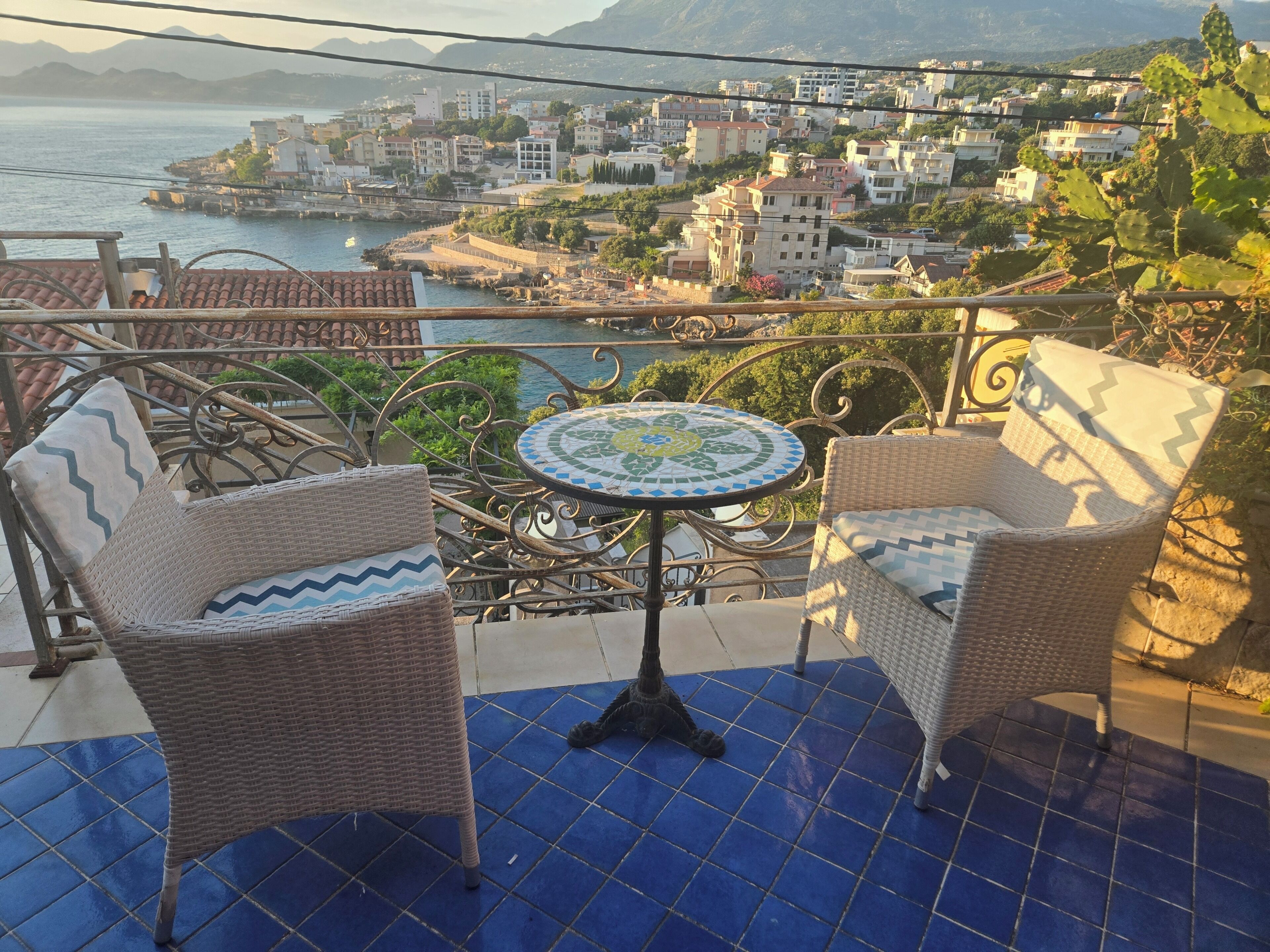 A balcony with wicker furniture and a tiled floor, overlooking a coastal town with buildings and a body of water.