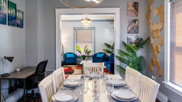 A dining area with a long table set for six, chairs, a lamp, and a view into a living room with blue chairs and plants. - Spacious 4BR Home Near Niagara Falls State Park | Sleeps 10 | Pet-Friendly (Niagara Falls)