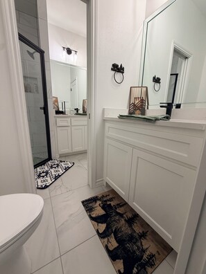 Een badkamer met witte meubels, een grote spiegel, een douche en een vloerkleed met een patroon.