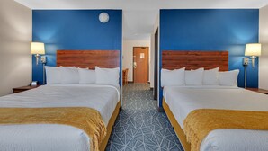 A hotel room with two beds, a wooden headboard, a patterned carpet, and a nightstand with a lamp.