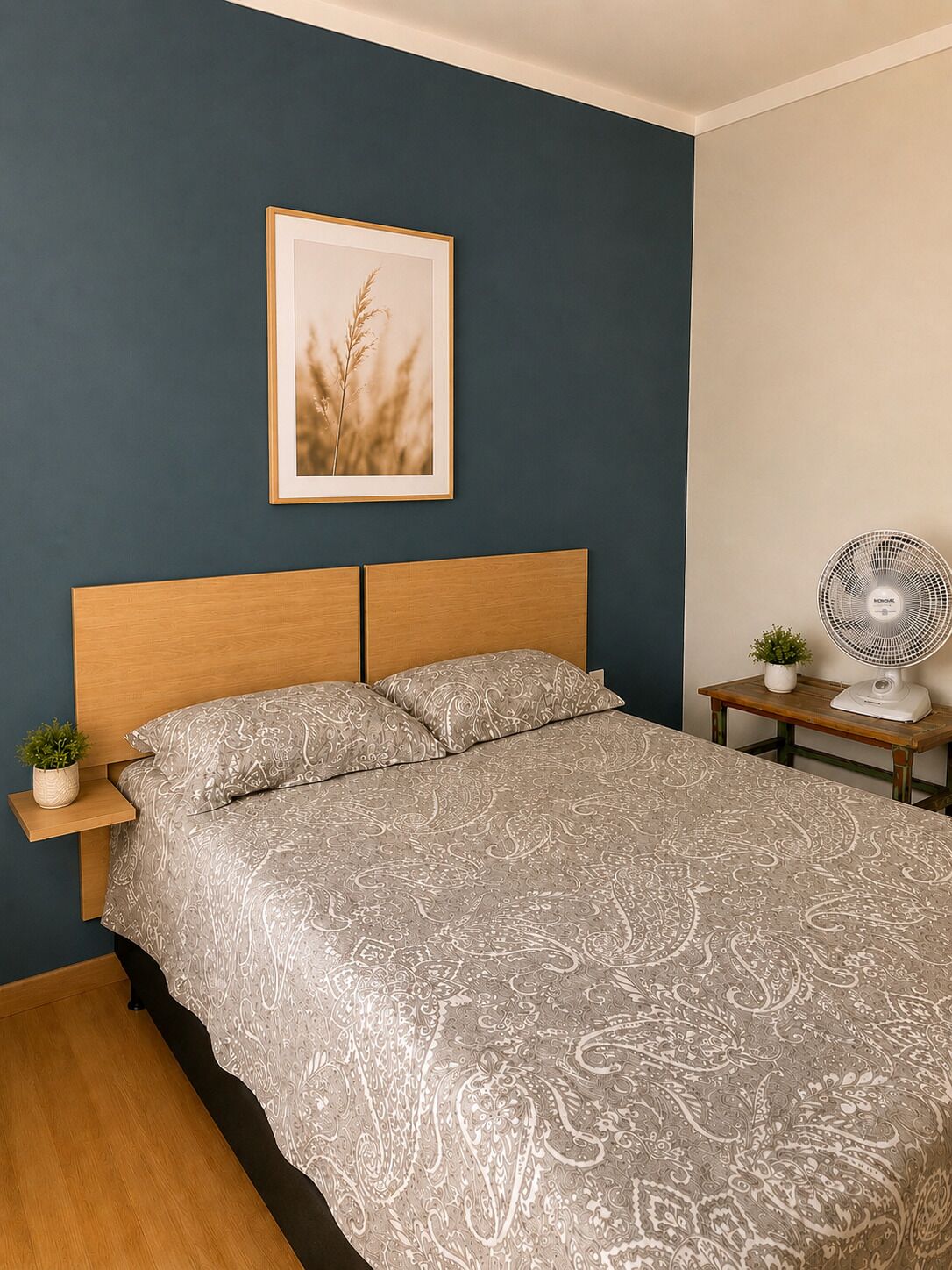 Une chambre à coucher avec un lit, une table de chevet ornée d’une plante, un tableau encadré accroché au mur et un ventilateur sur pied.