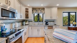 A kitchen with white cabinets, a stainless steel microwave, an oven, a sink, and a window with a view of greenery. - Charming pool home in Sea Oats, Sanibel Island (Sanibel)