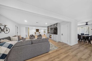 Una sala de estar moderna con un sofá gris, suelo de madera, televisor y un comedor con mesa y sillas.