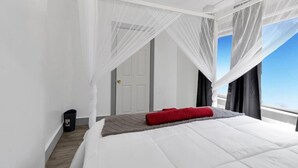 Un dormitorio con cama, cortinas blancas, almohada roja y vista al cielo.