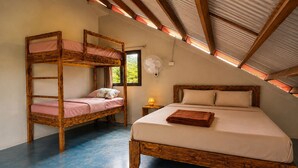 A room with a bunk bed and a single bed, both made of wood, with a fan on the wall and a window providing natural light.