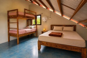Una habitació amb un llit superposat i un llit individual, tots dos de fusta, amb un ventilador a la paret i una finestra que deixa entrar llum natural.