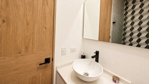 Un bany amb un armari de fusta, un lavabo blanc amb un recipient, un aixeta negra i un mirall que reflecteix una zona de banyera amb un disseny de motius geomètrics.
