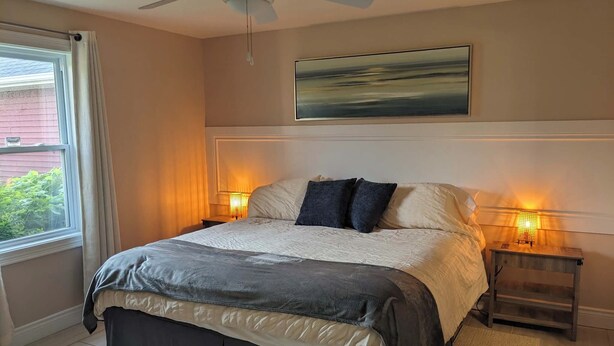 A bedroom with a bed, a ceiling fan, two bedside lamps, a window with curtains, and a painting on the wall.