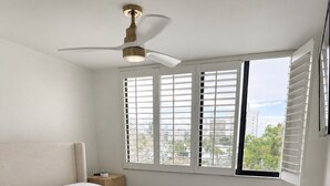 Un dormitorio con cama, ventilador de techo, ventana con persianas y vista a edificios.