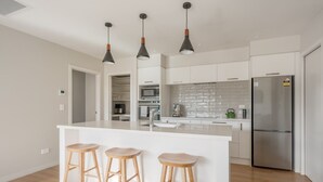 Une cuisine moderne dotée d’un îlot central, de trois tabourets en bois, d’appareils électroménagers en acier inoxydable et de luminaires suspendus.