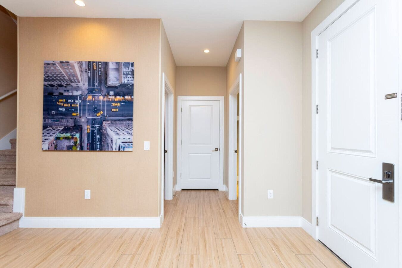 A hallway with a cityscape painting, wooden flooring, white doors, and a staircase on the left.