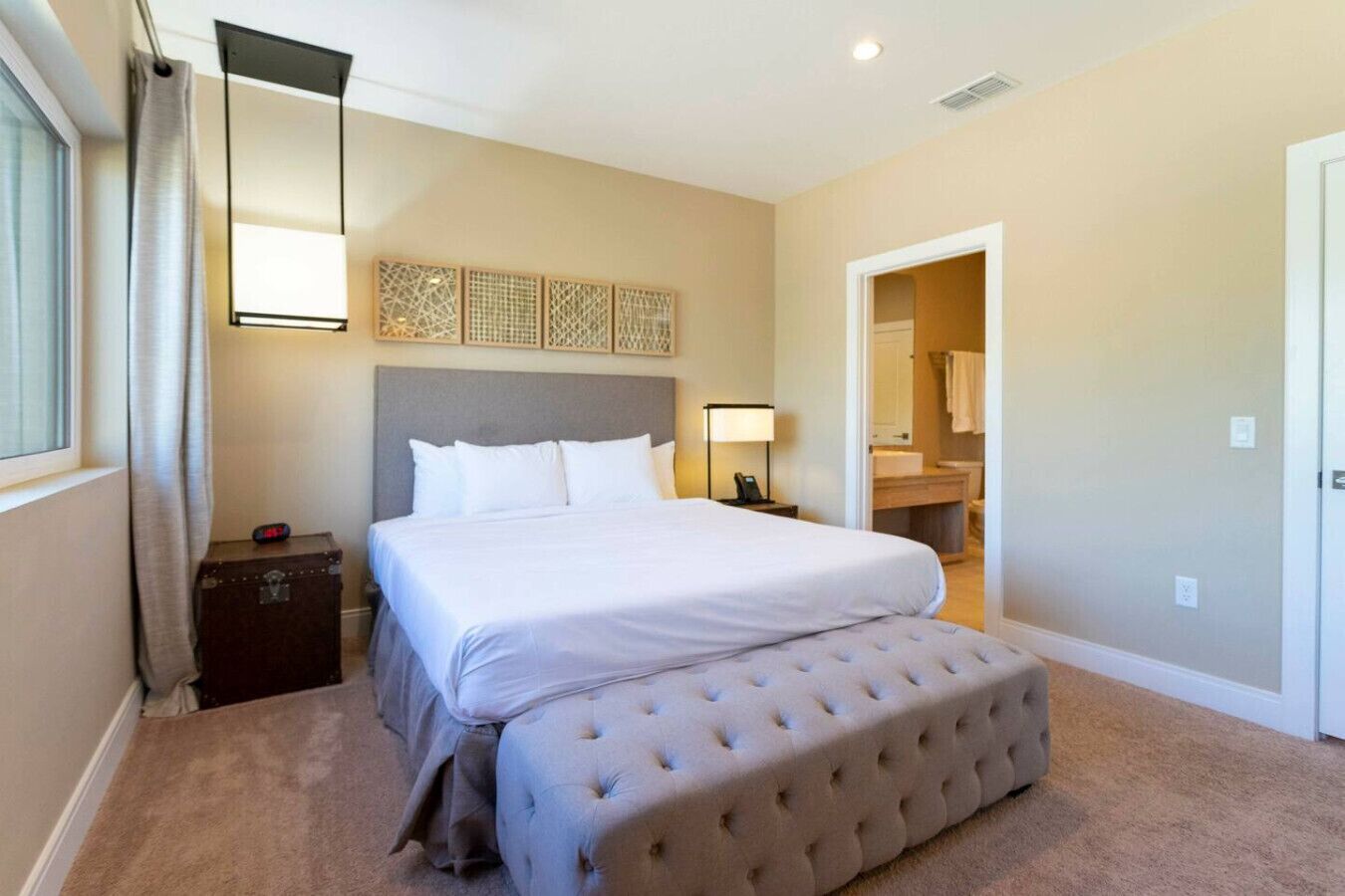 Camera d'albergo con un letto, un comodino, una lampada, una panca, un armadio e un bagno visibile attraverso una porta aperta.