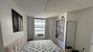 Ein Schlafzimmer mit geblattetem Bett, einem Fenster mit Vorhängen, einem großen abstrakten Gemälde und einem Spiegel.