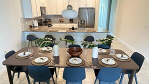 Een moderne keuken met witte kasten, een centraal eiland en een eethoek met blauwe stoelen en een bruine tafel.