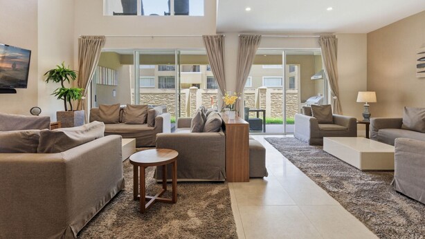 Ein großzügiger Wohnbereich mit hoher Decke, großen Fenstern und Blick ins Freie. Es gibt verschiedene Sitzgelegenheiten, darunter Sofas und ein Couchtisch.
