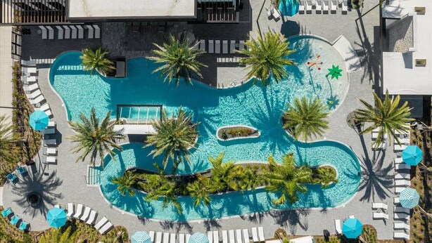 Vue aérienne d’un complexe hôtelier avec une grande piscine centrale, entourée de palmiers et de chaises longues.