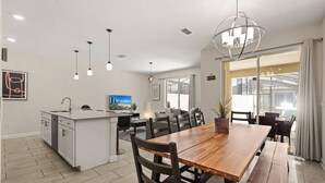 Cocina moderna con una isla central, comedor con mesa y sillas de madera, iluminación colgante y ventanas amplias.