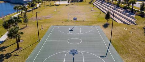 Una cancha de baloncesto exterior con aro, rodeada de césped y palmeras, con casas al fondo.