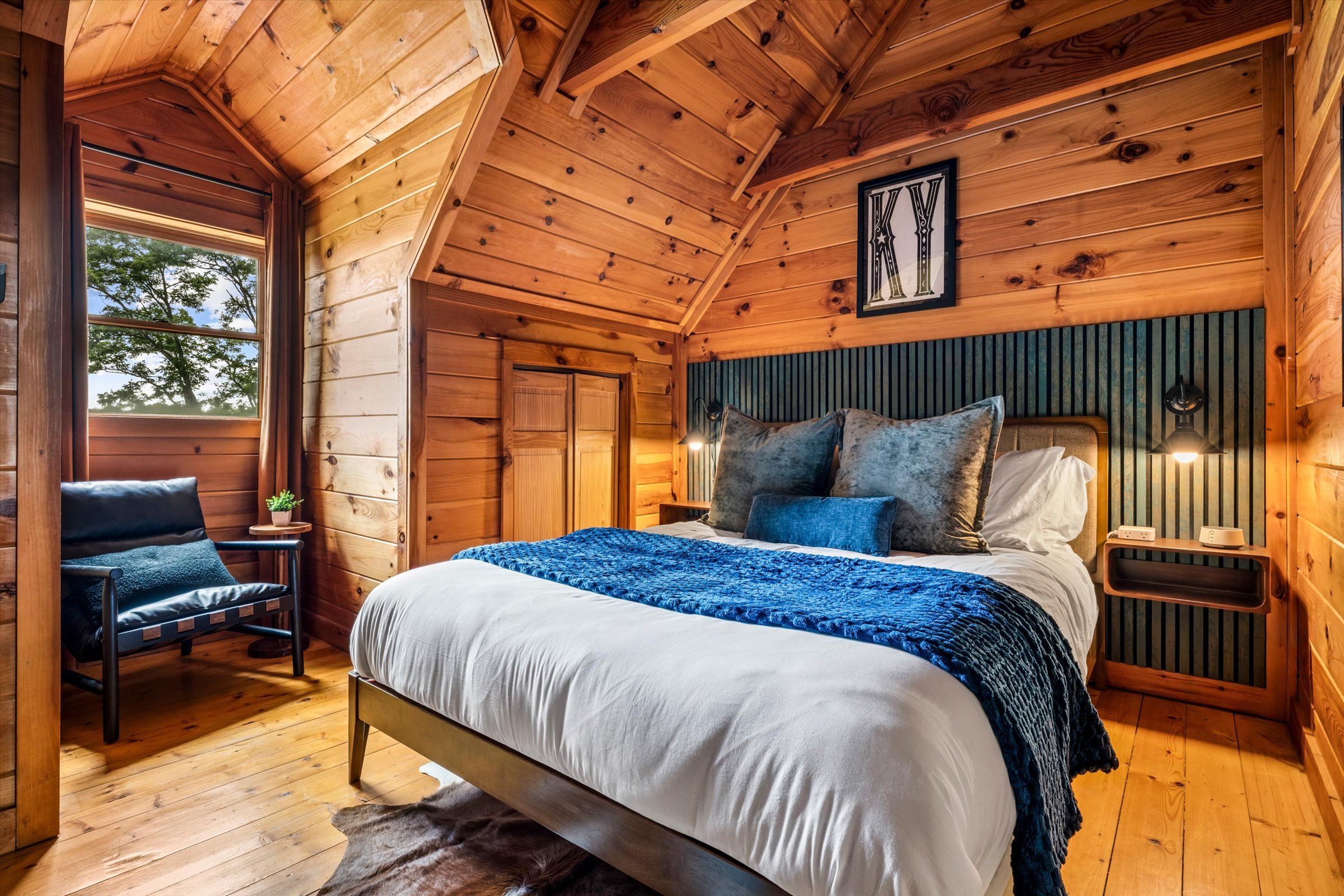 Ein Schlafzimmer einer Holzhütte mit einem Bett, einem Sessel, einem Nachttisch und einem Fenster.
