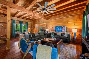 A spacious living room with a high ceiling, wooden beams, a ceiling fan, and a large painting.
