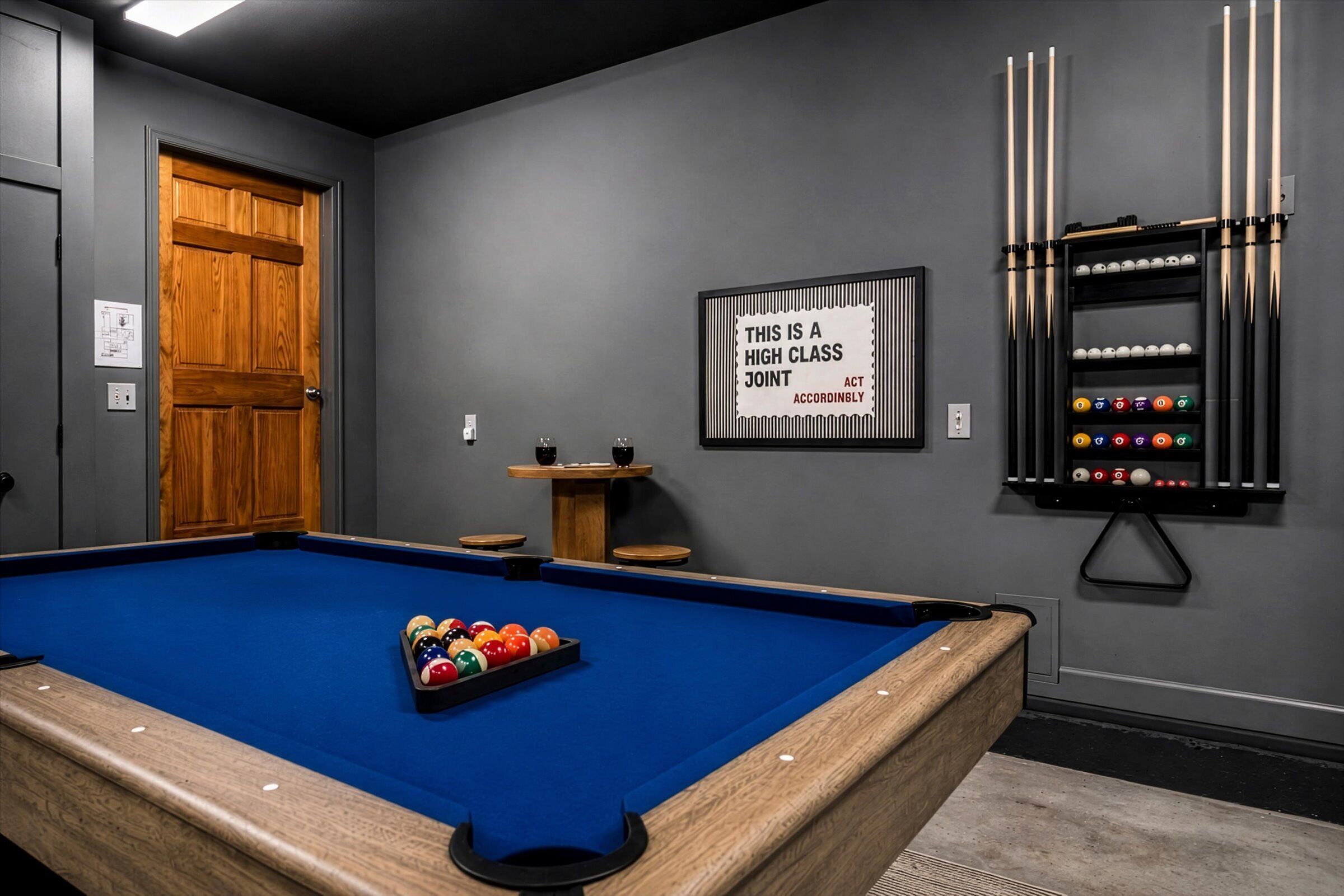 Una mesa de billar con bolas, un soporte para tacos, y un cartel que dice 'THIS IS A HIGH CLASS JOINT ACT ACCORDINABLY' en una habitación con paredes grises y una puerta de madera.