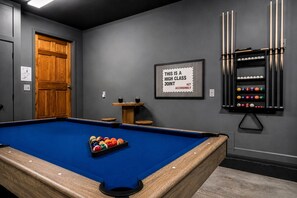Een pooltafel met ballen, een biljarthouder en een bord met de tekst 'THIS IS A HIGH CLASS JOINT ACT ACCORDINABLY' in een ruimte met grijze wanden en een houten deur.