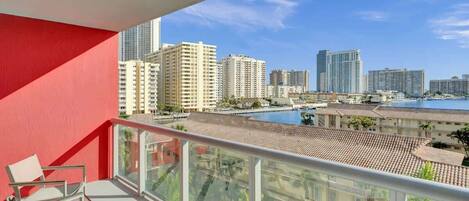 A balcony with a red wall, a glass railing, and a view of high-rise buildings and a body of water.