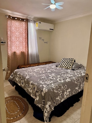 Un dormitorio con cama, ventilador de techo, equipo de aire acondicionado, pared con una frase motivadora y una alfombra estampada.