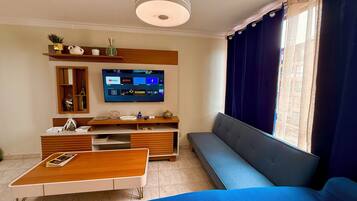 Ett vardagsrum med en platt-TV, en soffa, ett soffbord och ett fönster med gardiner.