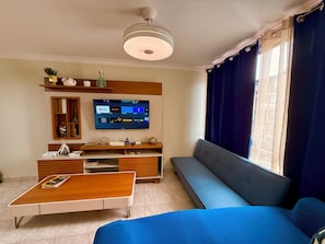 Una sala de estar con una televisión de pantalla plana, un sofá, una mesa de centro y un ventanal con cortinas.