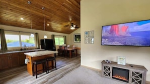 A modern kitchen with a wooden ceiling, a large island with stools, a dining area with chairs, a flat-screen TV mounted on the wall, and a fireplace. - Hawaii's Trivia Hale an Off the Beaten Path Oasis | AC | Family Friendly (Mountain View)