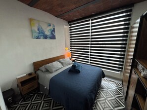 Une chambre à coucher avec un lit, une table de chevet, une lampe, un tableau au mur et une fenêtre avec des stores.
