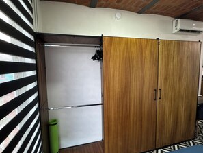 Ein Zimmer mit einem schiebbaren Holzschrank, einer weißen Tür, einem grünen Mülleimer und einem gestreiften Fensterrollo.