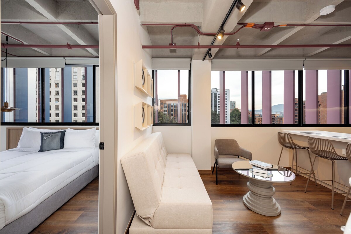 Quarto de hotel moderno com cama, sofá, área de jantar com mesa e cadeiras, e amplas janelas com vista para a cidade.