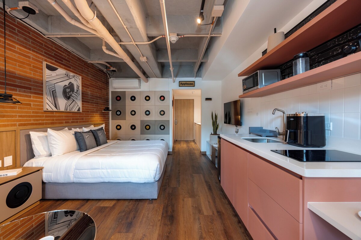 A modern hotel room with a large bed, a kitchen area with a sink and countertop, and a brick wall with a framed picture.