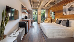 Una habitación de hotel moderna con una cama grande, un escritorio con silla, una guitarra, un televisor y vistas a zonas verdes por la ventana.