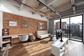 Una sala moderna con pared de ladrillo, un sofá blanco, un escritorio con una silla, una guitarra y un ventanal con vistas a edificios.