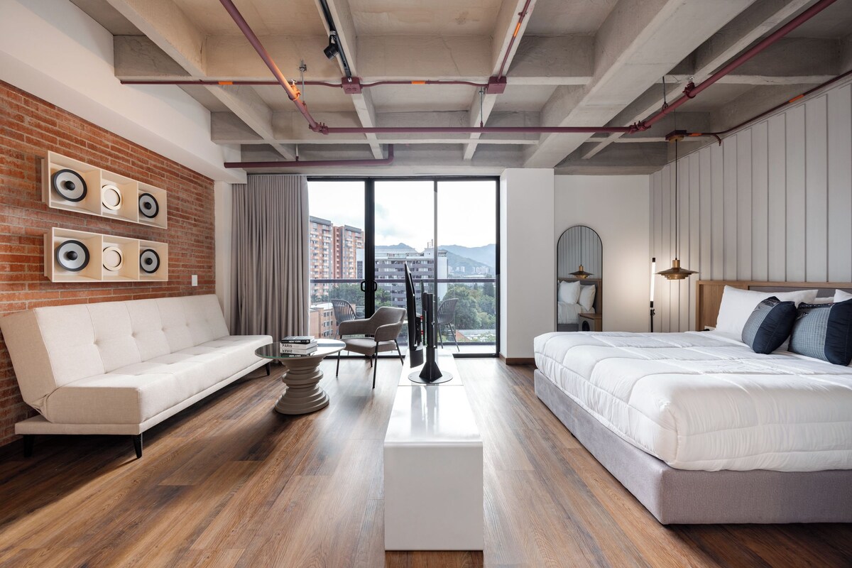 Quarto de hotel moderno com parede de destaque em tijolo, sofá branco, cama com roupa de cama branca, mesa redonda e vista para edifícios por grandes janelas.