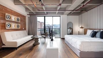 A modern hotel room with a brick accent wall, a white sofa, a bed with white bedding, a round table, and a view of buildings through large windows.