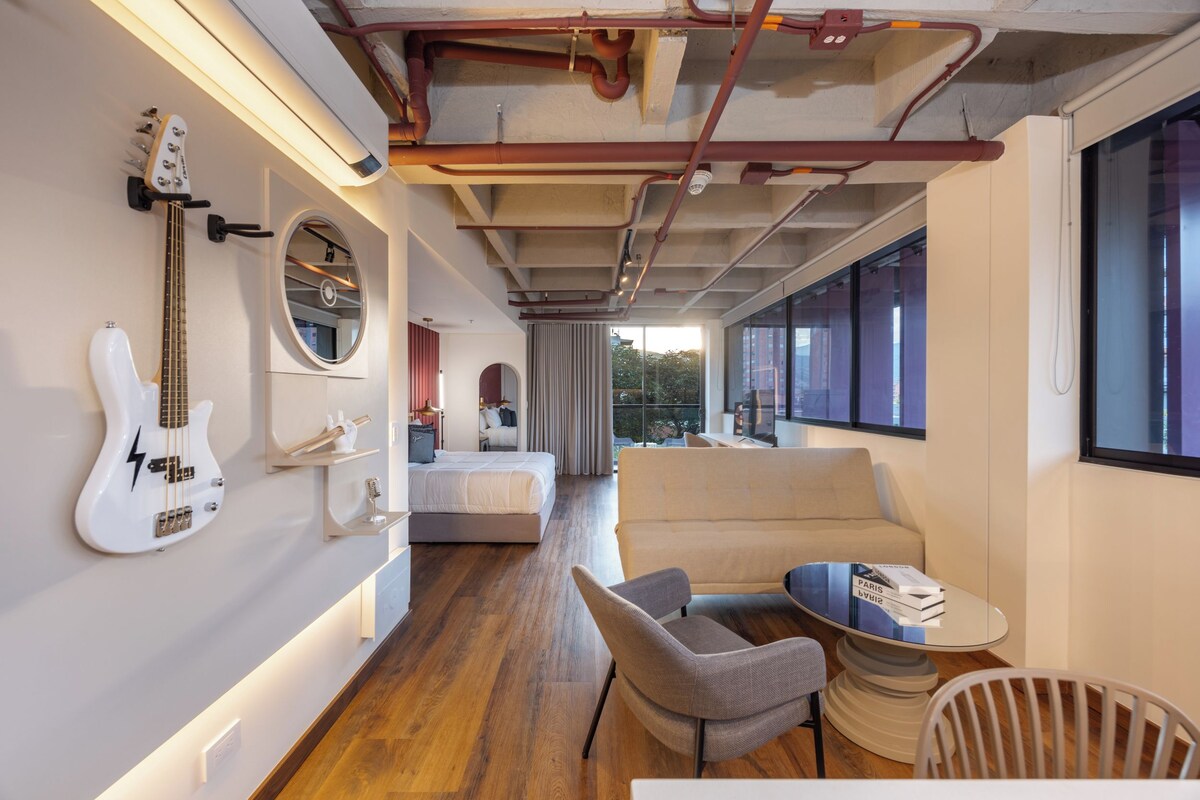 Quarto de hotel moderno com uma cama, área de estar com sofá e cadeiras, uma guitarra na parede e vista para o exterior.