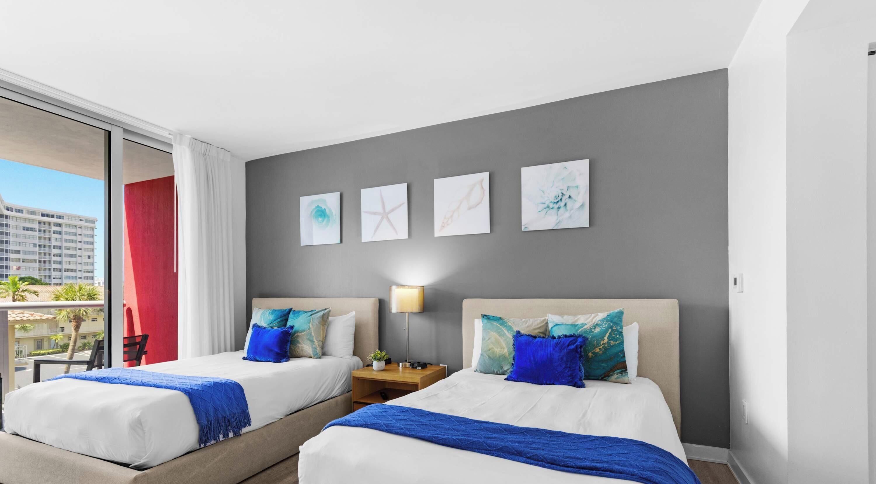 A hotel room with two beds, a nightstand, a lamp, and artwork on the wall.