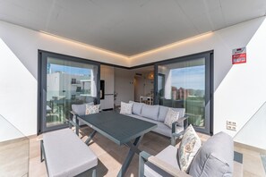 Ein moderner Außenbereich mit einer Sitzecke, Tisch und Stühlen, großen Glasfenstern und einer durch die Fenster einsehbarer Küche.
