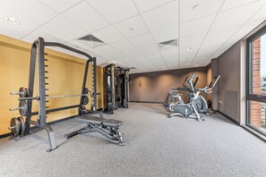 Ein Fitnessraum mit unterschiedlichen Trainingsgeräten, darunter eine Langhantelbank, ein Squat-Gestell und ein Crosstrainer.