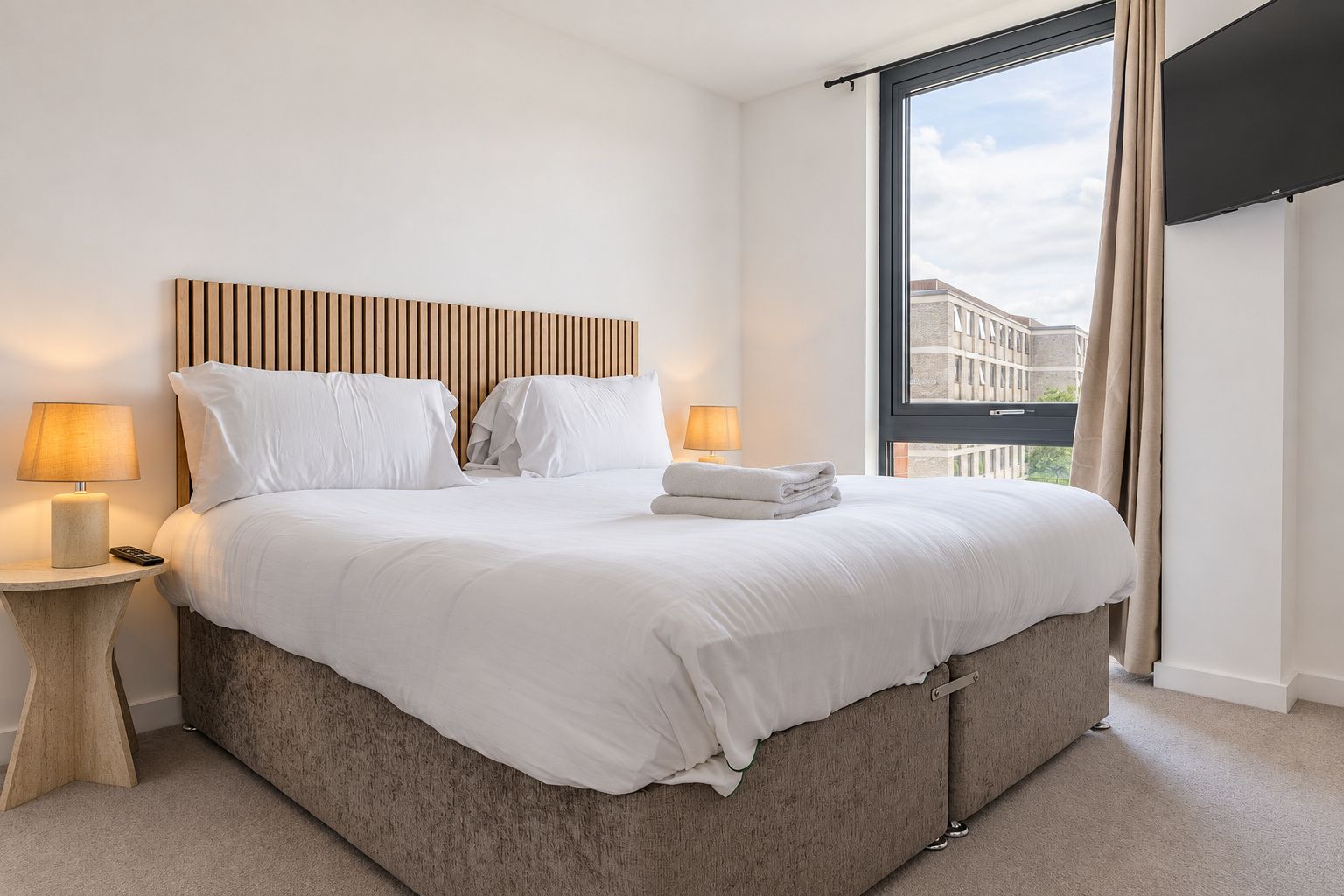 A hotel room with a large bed, two bedside tables with lamps, a window with a view of buildings, and a wooden headboard.