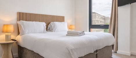 A hotel room with a large bed, two bedside tables with lamps, a window with a view of buildings, and a wooden headboard.