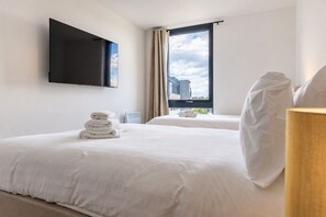 Hotellihuone, jossa on kaksi sänkyä, seinälle asennettu taulutelevisio, ikkuna, jossa on verhot, ja yöpöydällä oleva lamppu.