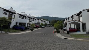 Une rue résidentielle bordée de maisons blanches, avec des voitures garées et un paysage vallonné en arrière-plan.