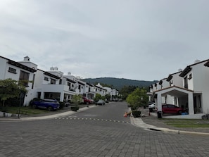 Una calle residencial con casas blancas, coches aparcados y un paisaje montañoso al fondo.