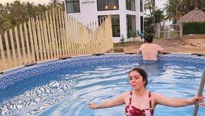 Una piscina con una valla de madera, un edificio con un letrero que dice 'JARDIN DEL MAR' y palmeras al fondo.