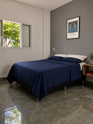 Une chambre à coucher avec un lit, une fenêtre avec des stores, un tableau encadré au mur et une table de chevet avec une plante.